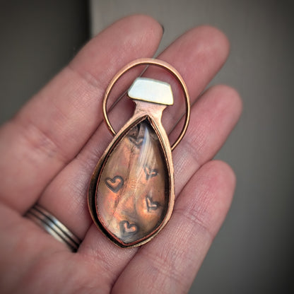 A copper pendant which looks like a potion bottle has stamped hearts under an almost clear rose quartz and a brass cap.