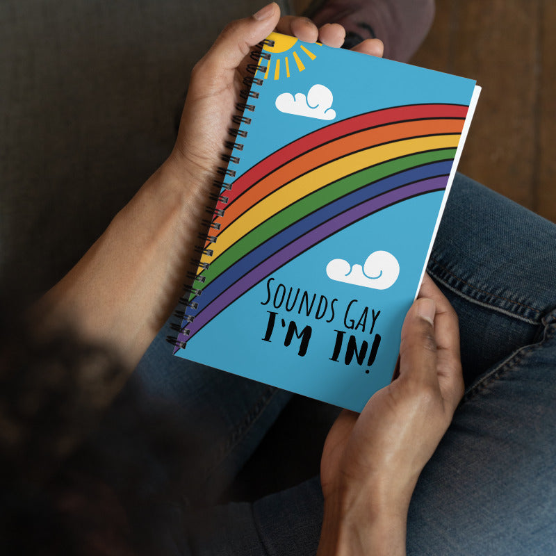 A model holds a spiral notebook with a bright rainbow across the front, a sun in the corner, and clouds in the sky has bold writing that says, "Sounds Gay, I'm in!"