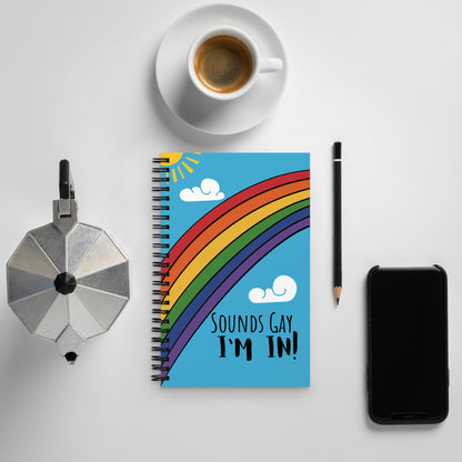 A spiral notebook with a bright rainbow across the front, a sun in the corner, and clouds in the sky has bold writing that says, "Sounds Gay, I'm in!"