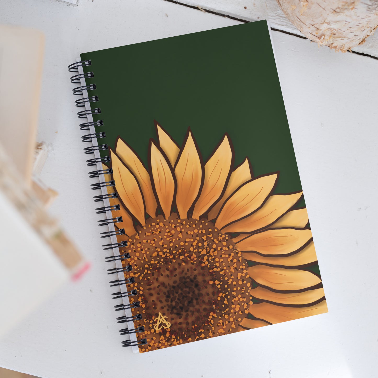 A spiral notebook has a large painted sunflower coming from one corner on a forest green background.