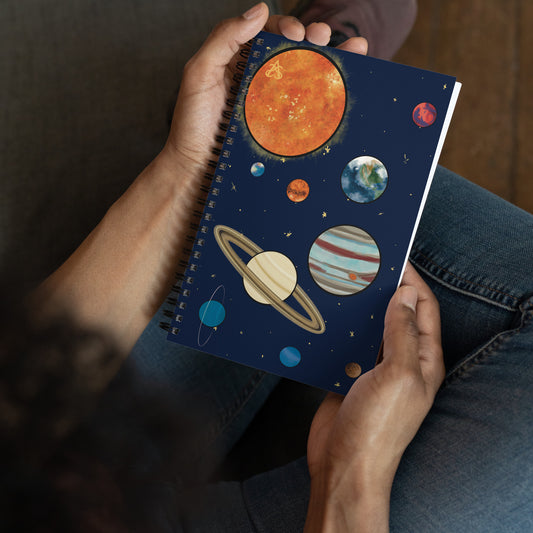 A pair of hands holds a spiral notebook with art of the solar system by Aras Sivad on the cover.
