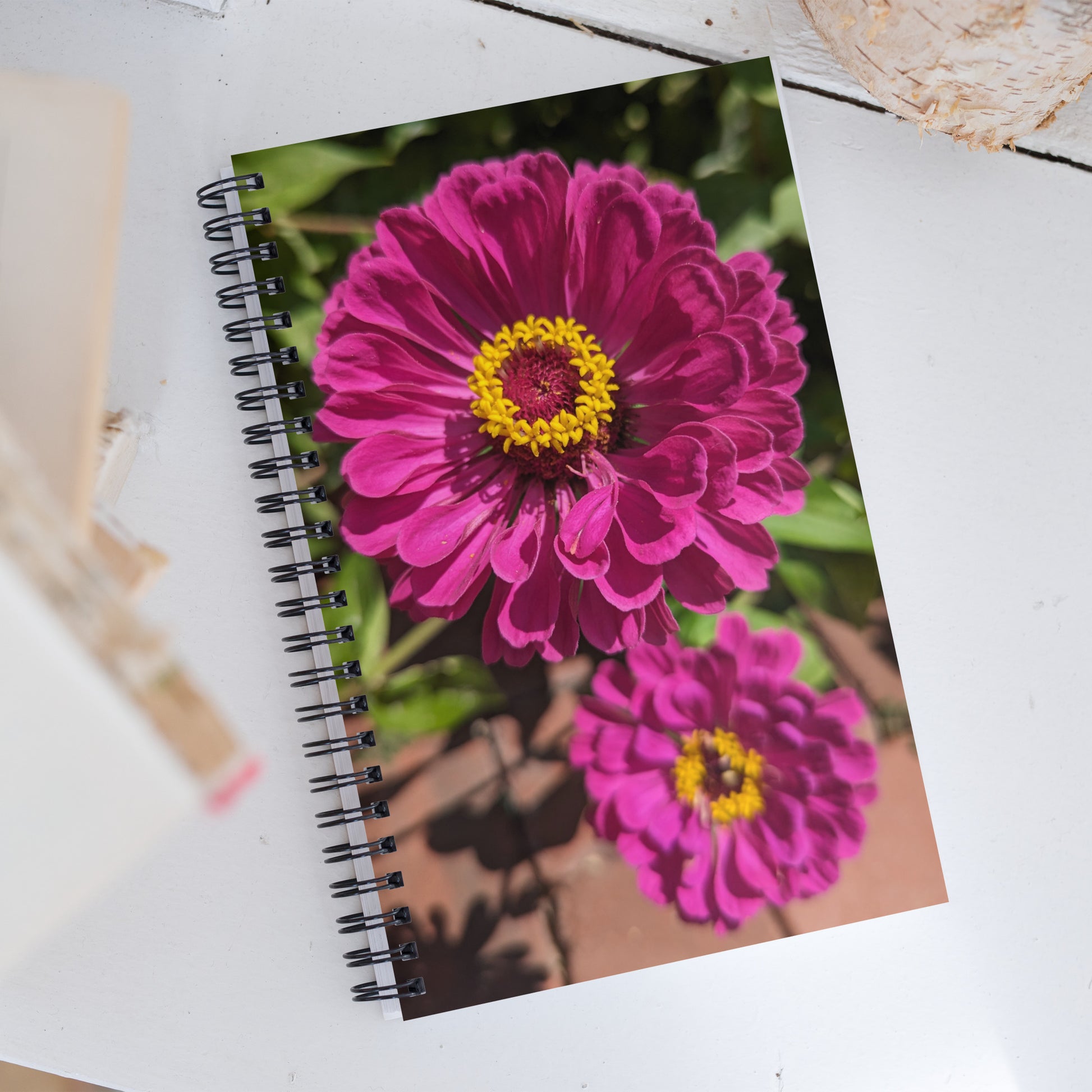 A spiral notebook is covered with a picture of a pair of bright fuchsia zinnias on a sunlit bush by Aras Sivad.