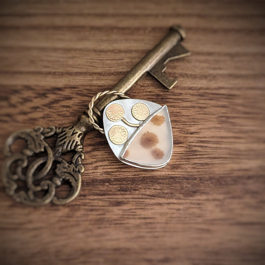 A silver pendant with gold brass flower stamped accents seems to grow out of the side of a bezel holding a rounded triangle shape scenic agate with flower-like inclusions.
