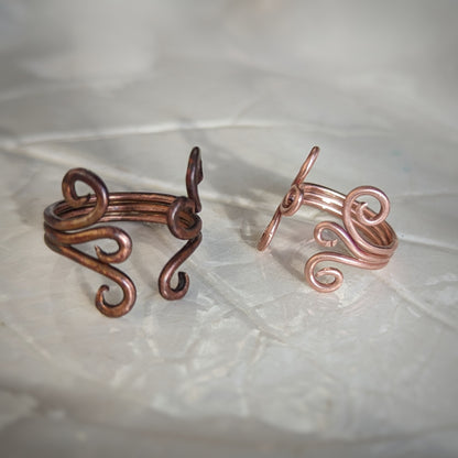 Two adjustable copper rings consist of three wires soldered together, each side ending in graceful swirls. One is has a rainbow appearing patina and the other is bright and shiny.