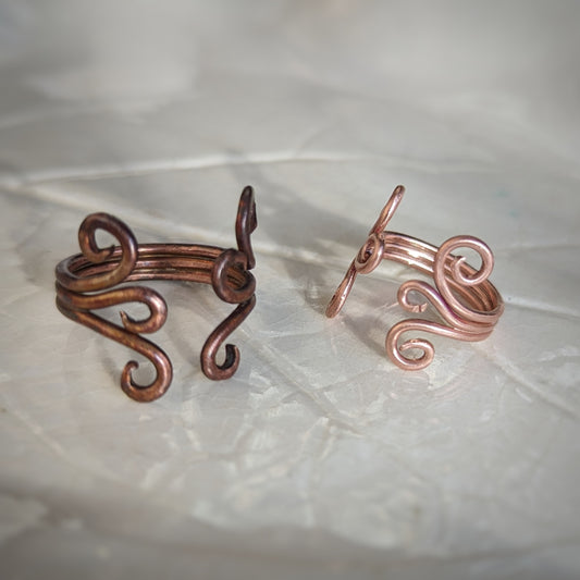 Two adjustable copper rings consist of three wires soldered together, each side ending in graceful swirls. One is has a rainbow appearing patina and the other is bright and shiny.