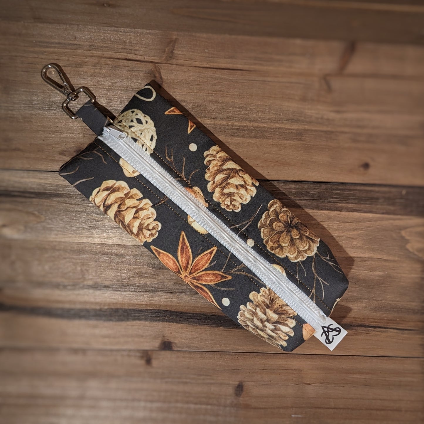 A long skinny pencil case with a keychain top has star anise, acorn, sticks, and pinecones outside and a white zipper up the middle.