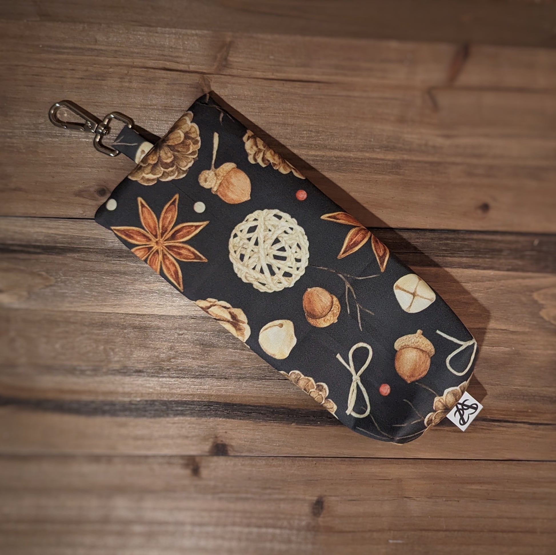 A long skinny pencil case with a keychain top has star anise, acorn, sticks, and pinecones outside is turned over to show more of the pattern on the back and the keychain clip flat top.