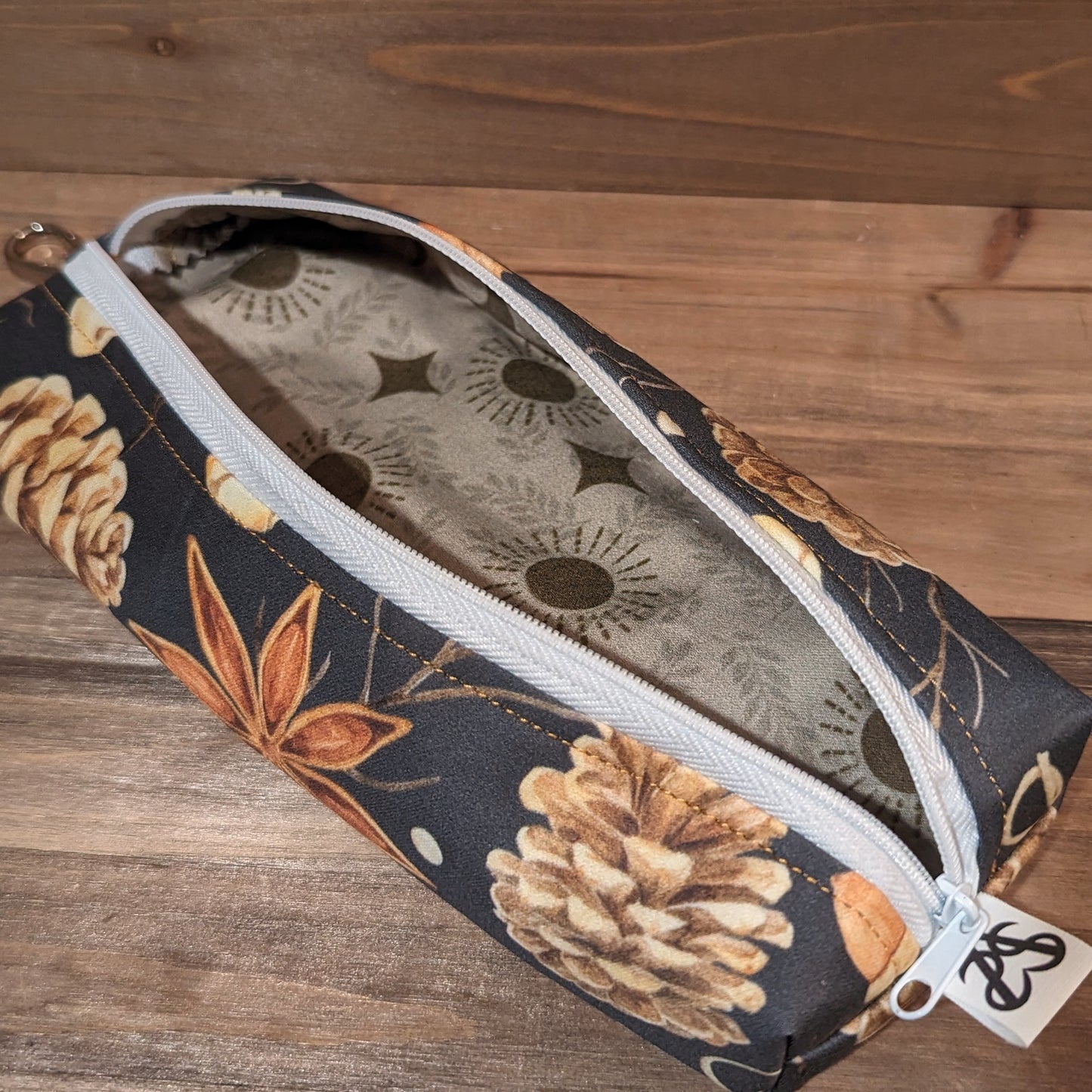 A long skinny pencil case with a keychain top has star anise, acorn, sticks, and pinecones outside and a white zipper up the middle open to show the moon, sun, and star print liner.
