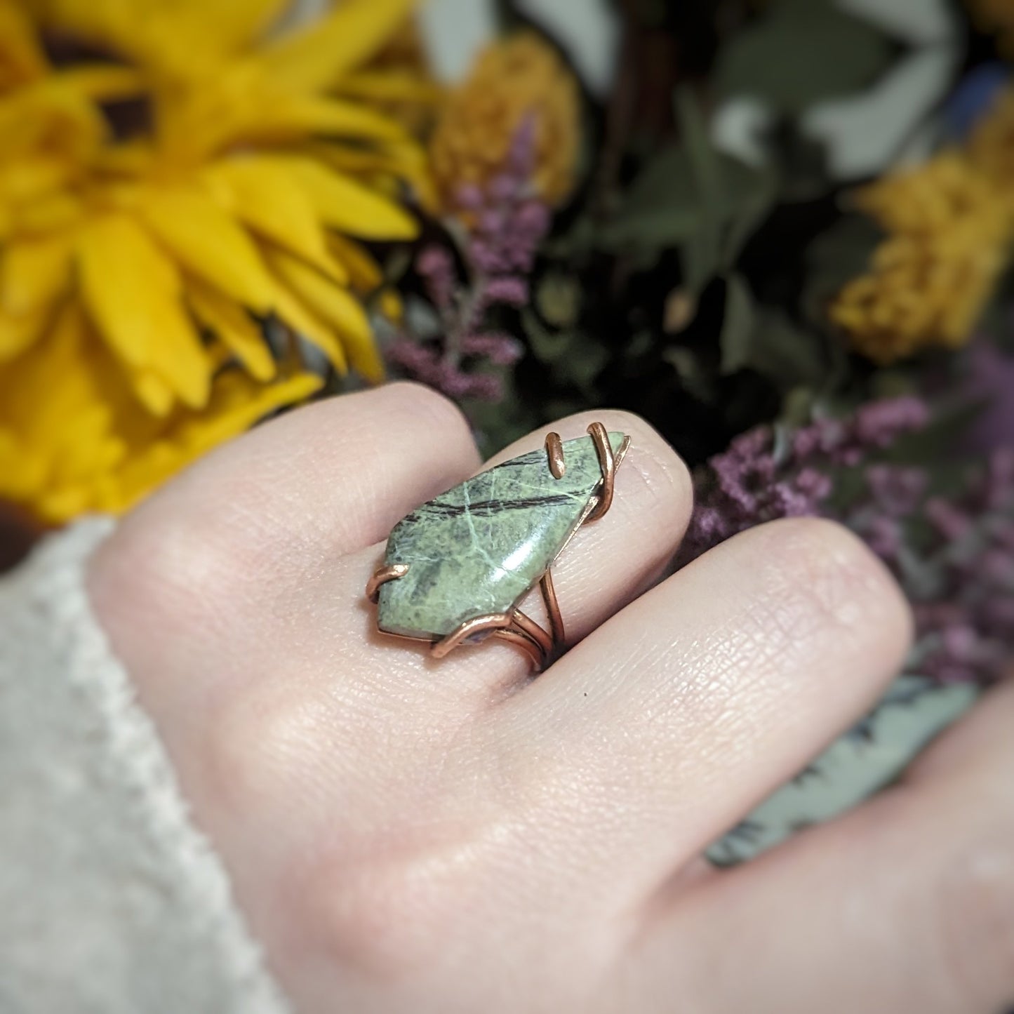 Aras models a copper ring with a skinny kite shaped green Atlantisite stone held on by swirling asymmetrical prongs.