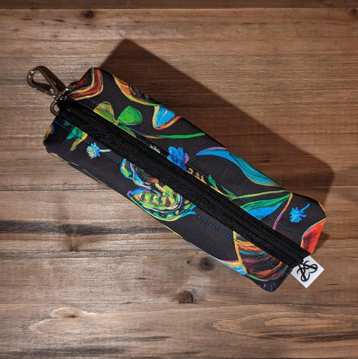 A long bag with box ends has a colorful forest pattern with pinecones, snails, frogs, acorns, florals, and keys, a black zipper down the middle, and a keychain clip on the top box shaped end.
