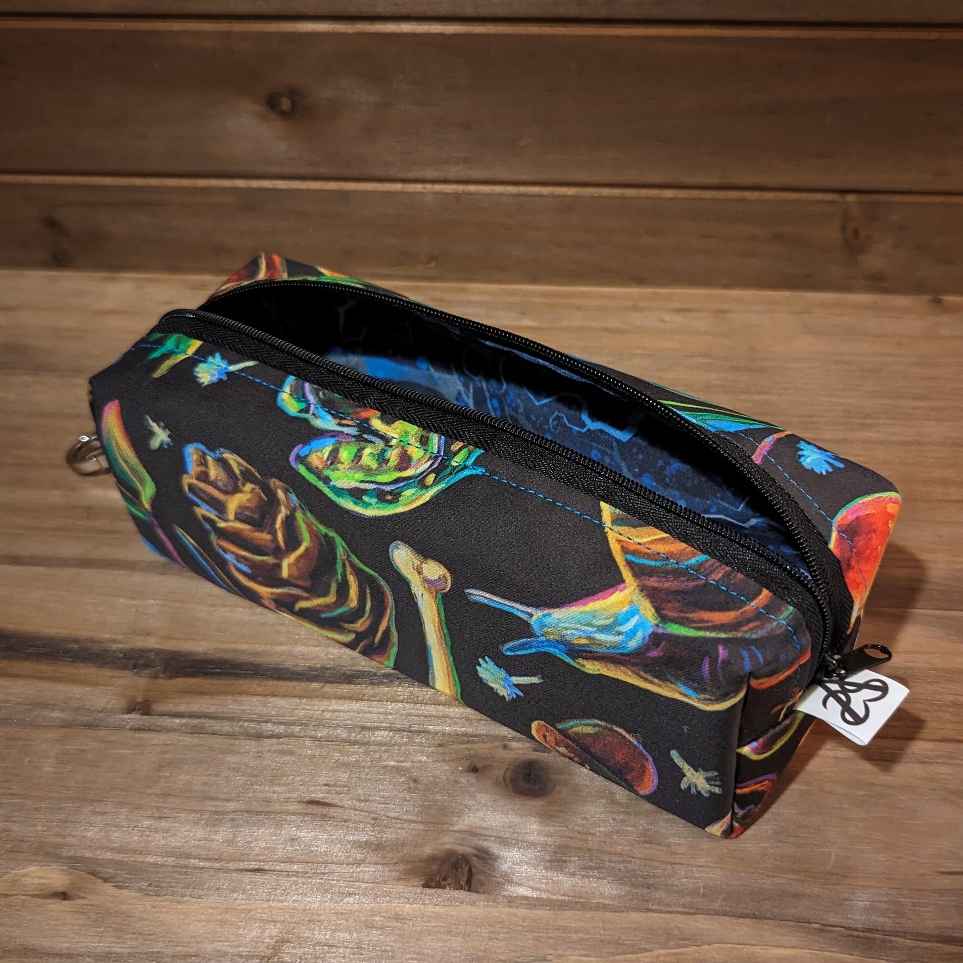 A long bag with box ends has a colorful forest pattern with pinecones, snails, frogs, acorns, florals, and keys, a black zipper down the middle open to show the blue spindly tree fabric inside, and a keychain clip on the top box shaped end.
