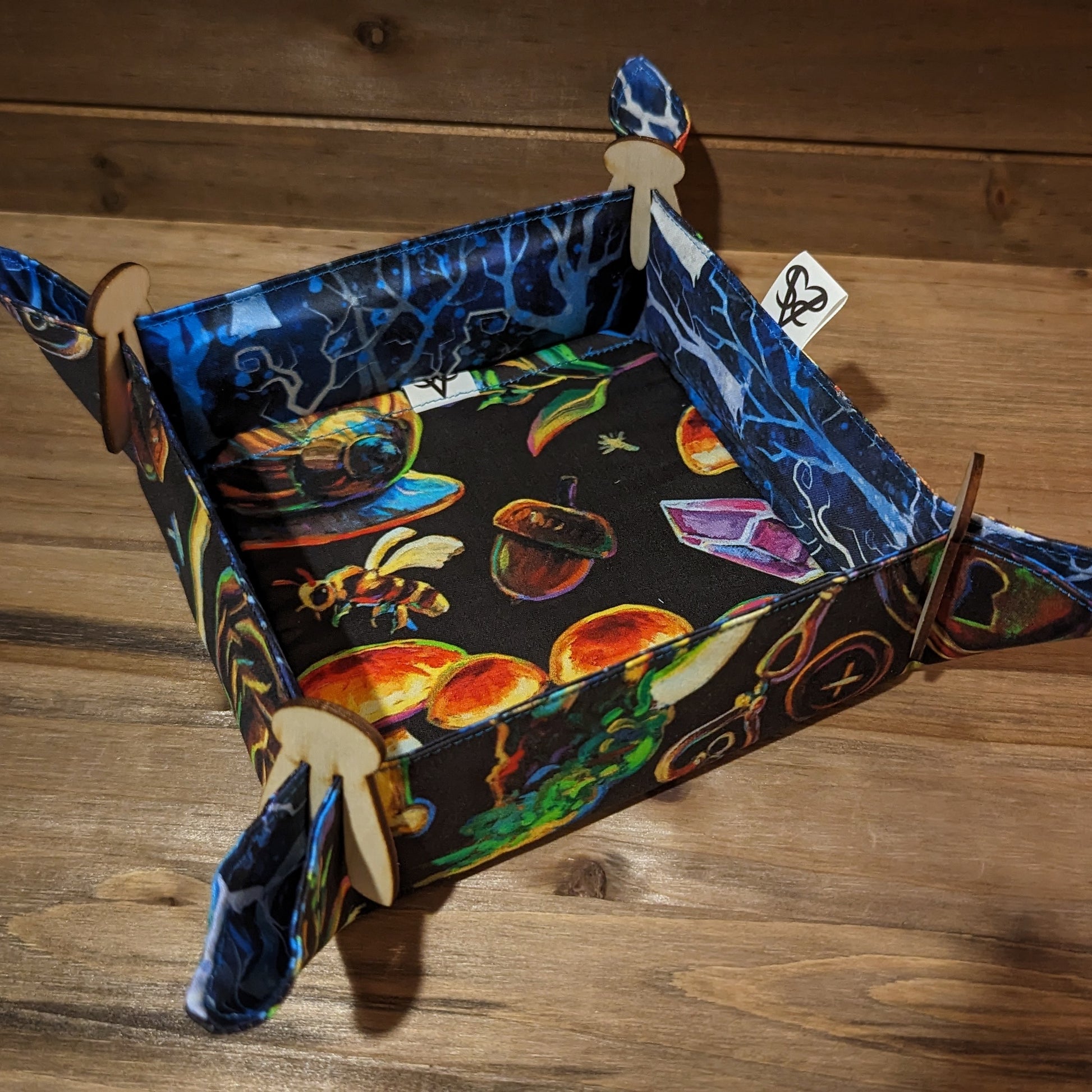 A dice tray with spindling trees in shades on blue on one side, colorful forest plants, bugs, and crystals on the other side, a matching insert in the bottom, and wood clips in the corners.