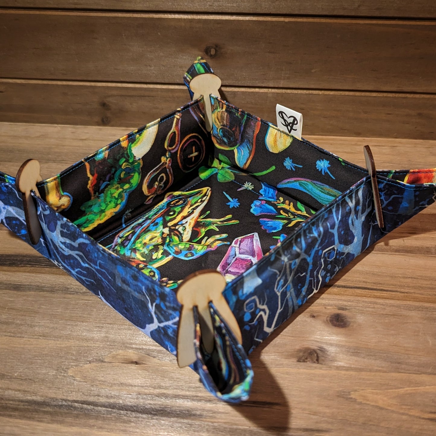 A dice tray with spindling trees in shades on blue on one side, colorful forest plants, bugs, and crystals on the other side, and wood clips in the corners.