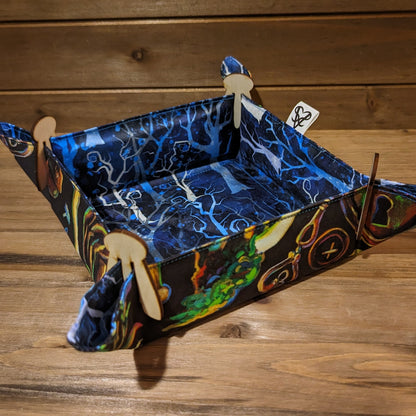 A dice tray with spindling trees in shades on blue on one side, colorful forest plants, bugs, and crystals on the other side, and wood clips in the corners.
