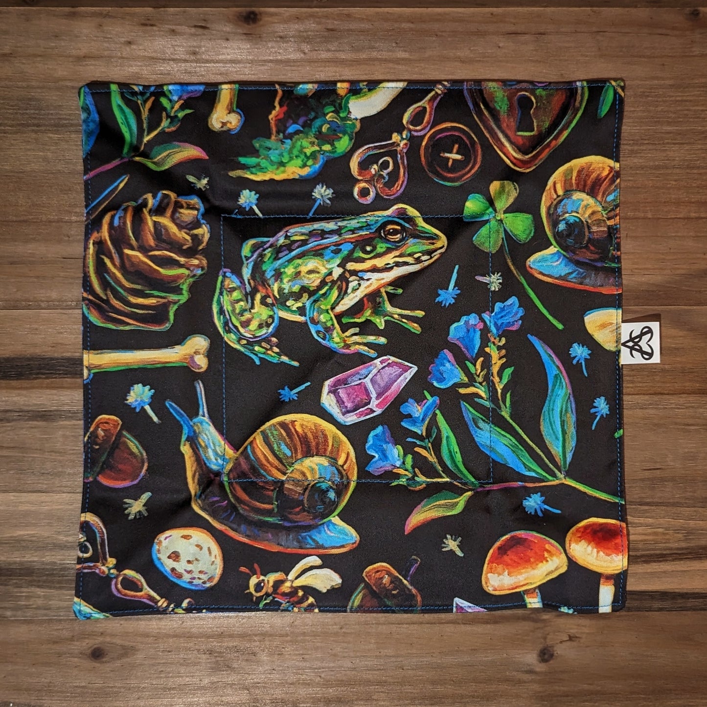 The witch's forest tray laid flat to show the entire colorful forest design on the first side with a frog, keys, bees, snails, crystals, mushrooms, pinecones, four leaf clovers, eggs, button, and blue flowers - accented with bright blue stitching.