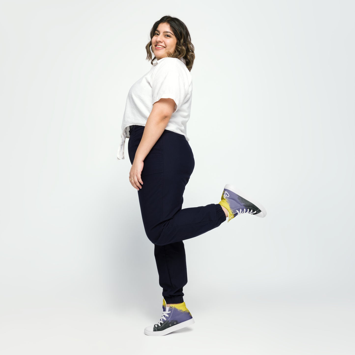A model wears a pair of high-top canvas shoes with cloudy color blocks of the yellow, purple, and black non-binary flag colors with white laces and accents, a white heart on the heel, and white Aras Sivad Studio logo on the tongue.
