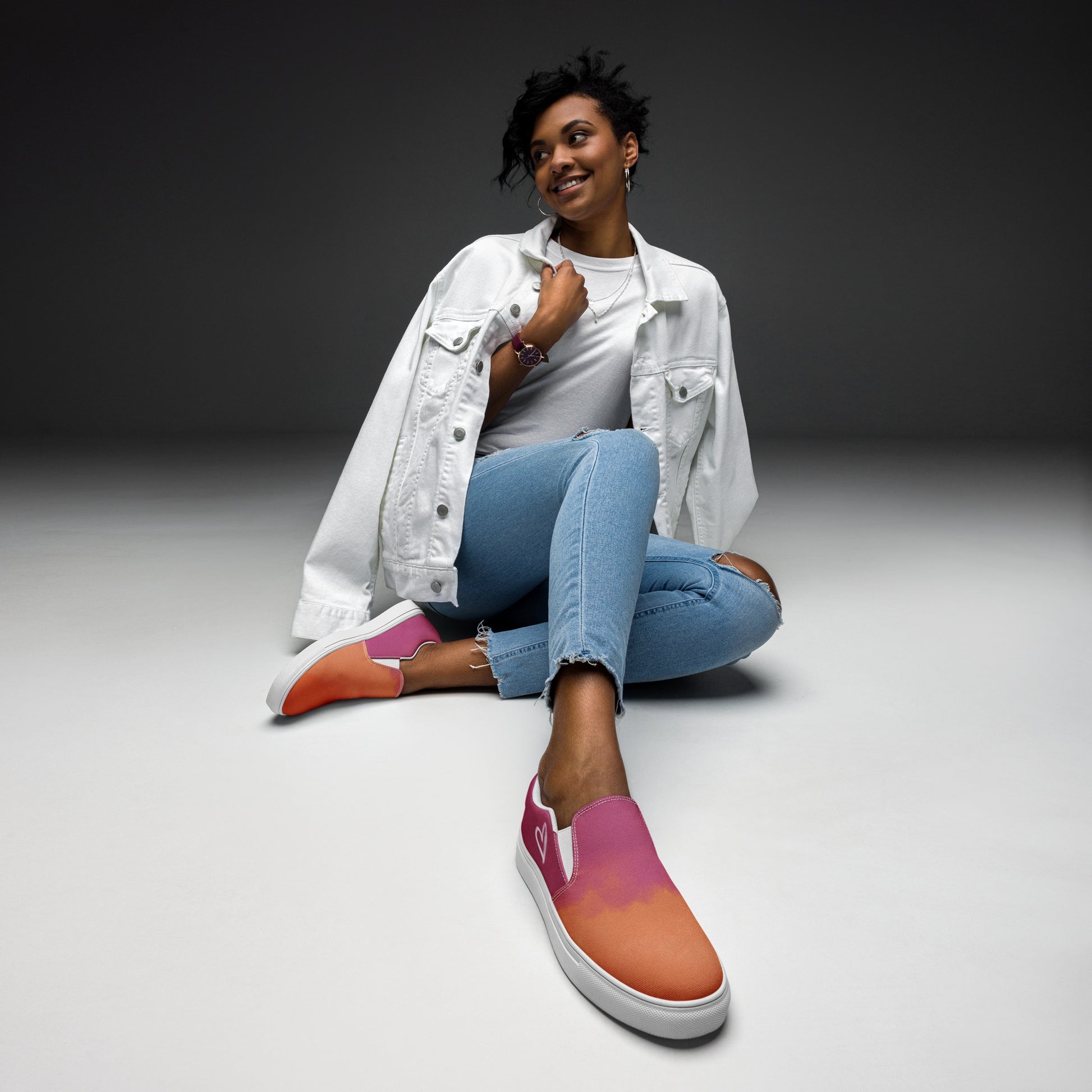 A model wears a pair of slip on canvas shoes with the colors of the lesbian pride flag in a cloudy texture, a white heart on the side, and the Aras Sivad logo on the back.
