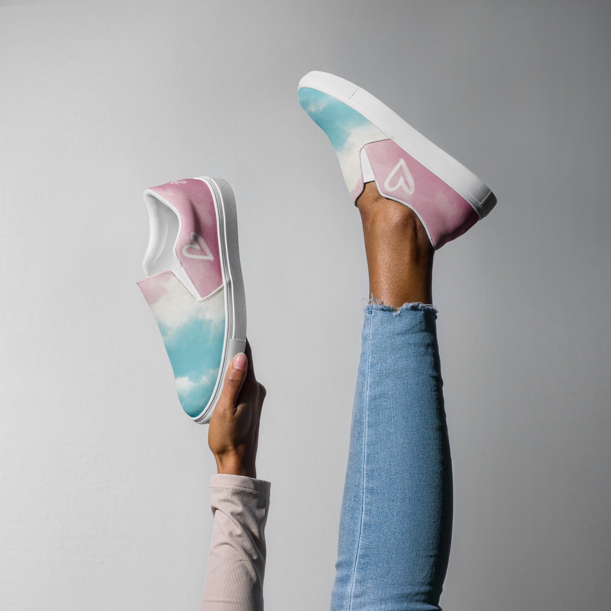 A model wears the Cloudy Transgender Pride flag slip on shoe on one foot and hold the match in the air for scale.