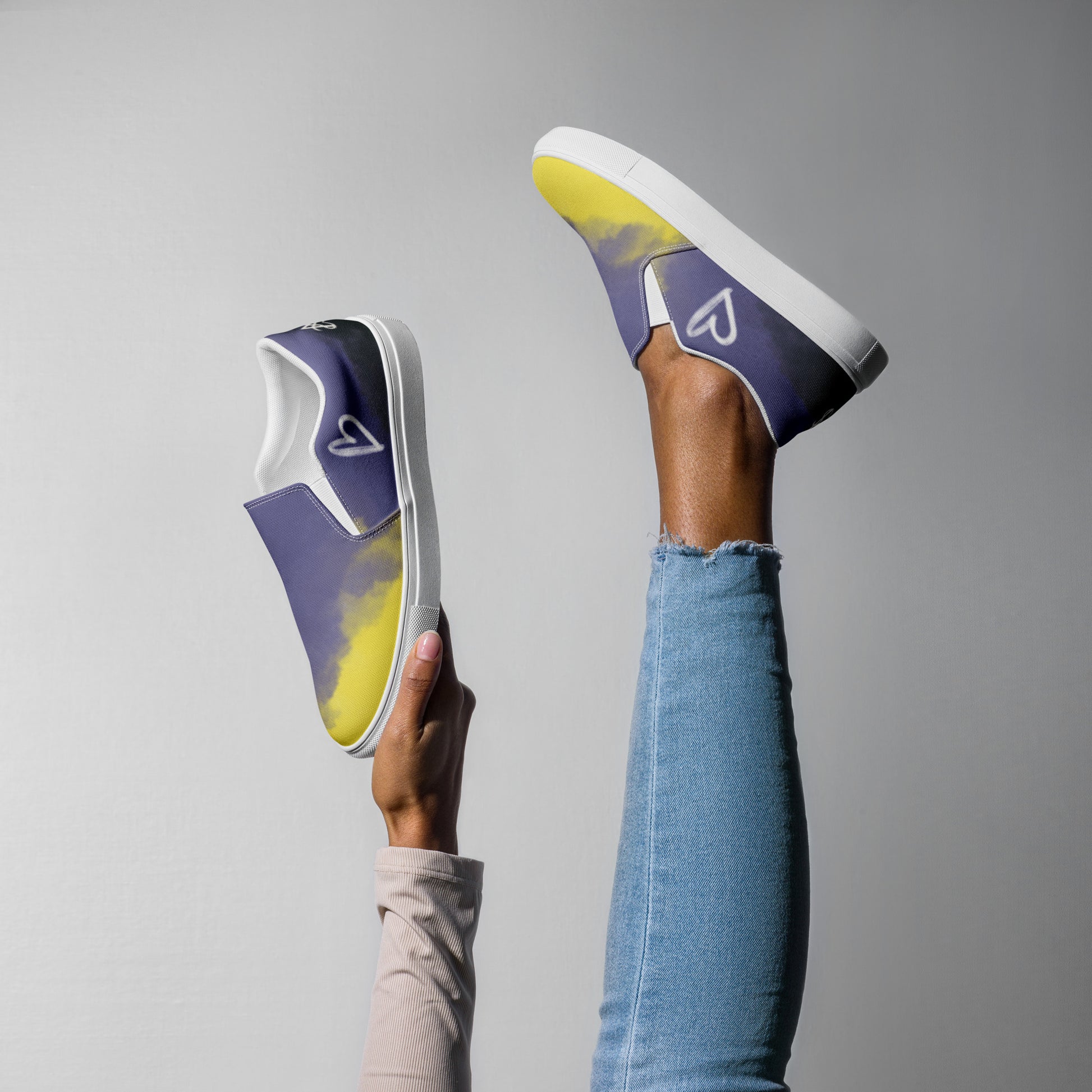 A model wears one and holds up a second slip-on shoe with the non-binary colors in wisps of clouds with a white hand drawn heart on the outside under the ankle and the Aras Sivad Studio logo in white on the back.