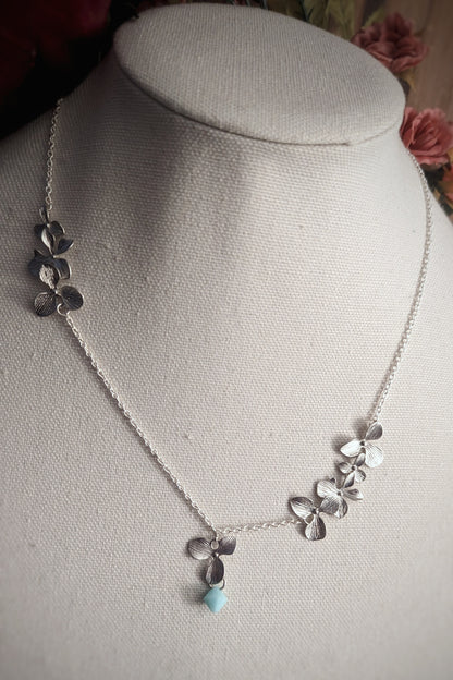 A fabric bust holds a dainty silver necklace with small sculpted orchid links suspended asymmetrically on a thin chain with a light blue drop hanging from a small silver orchid pendant.