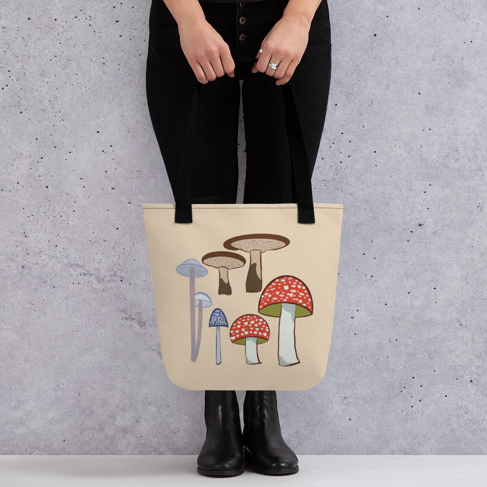 A model holds a tan canvas tote bag with black straps and illustrations of 4 different mushroom types on the front.