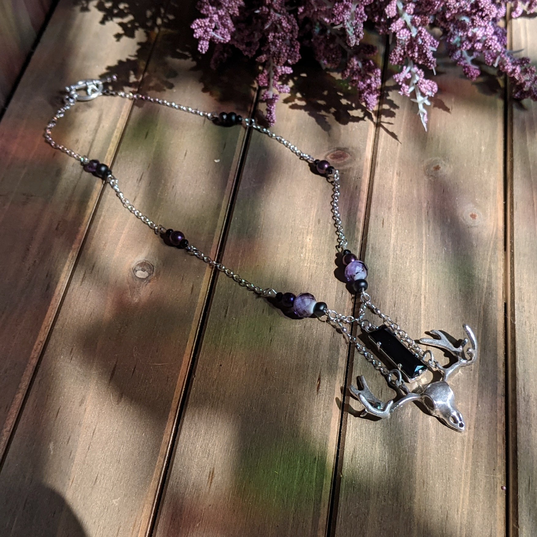 A silver deer skull necklace with a black rectangular stone hanging above it on a silver chain dotted with black and purple beads.
