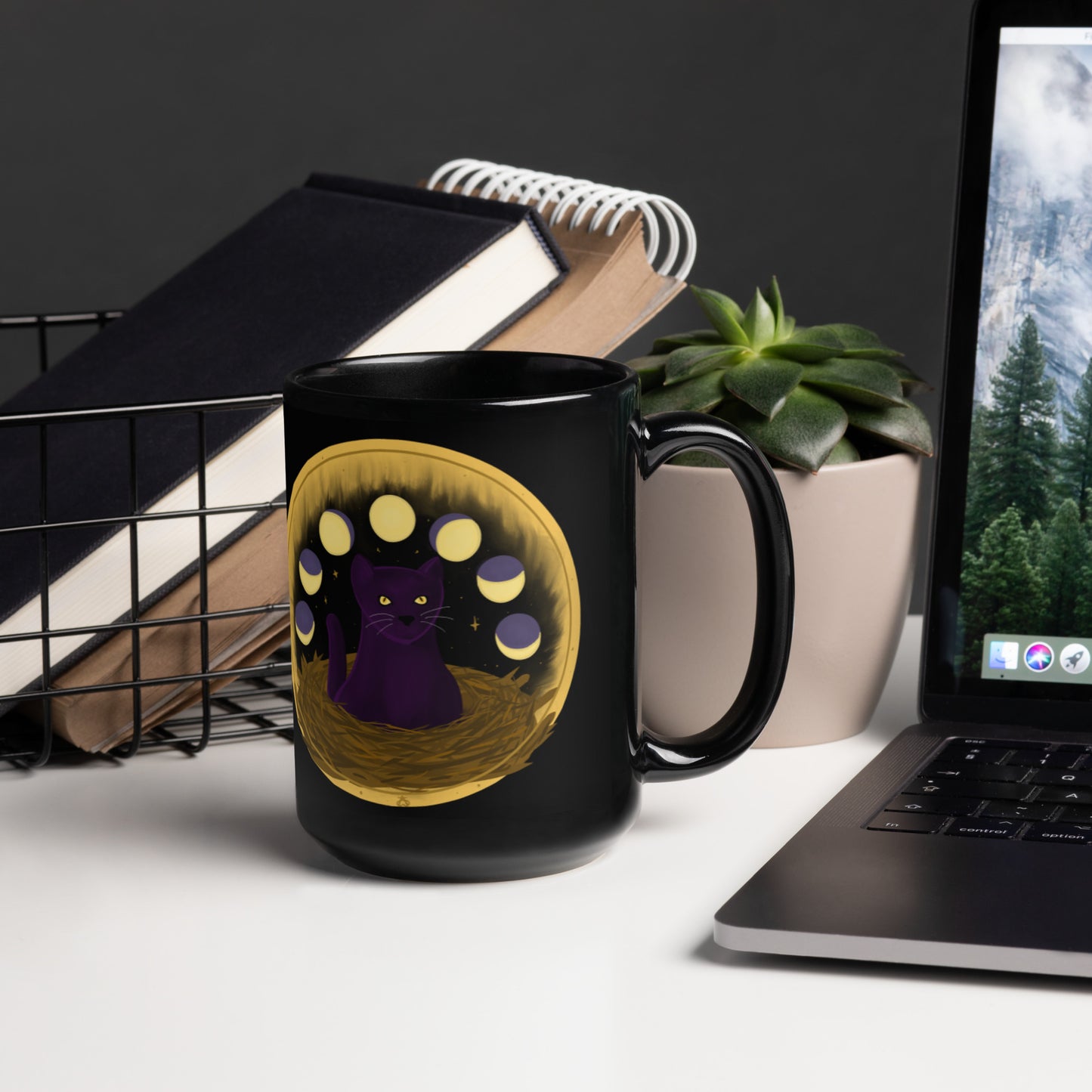 A black mug with a painting of a cat sitting in a nest with the phases of the moon over it, ringed in gold with twinkling gold stars behind it, sitting on a generic desk with a succulent, notebooks, and laptop.