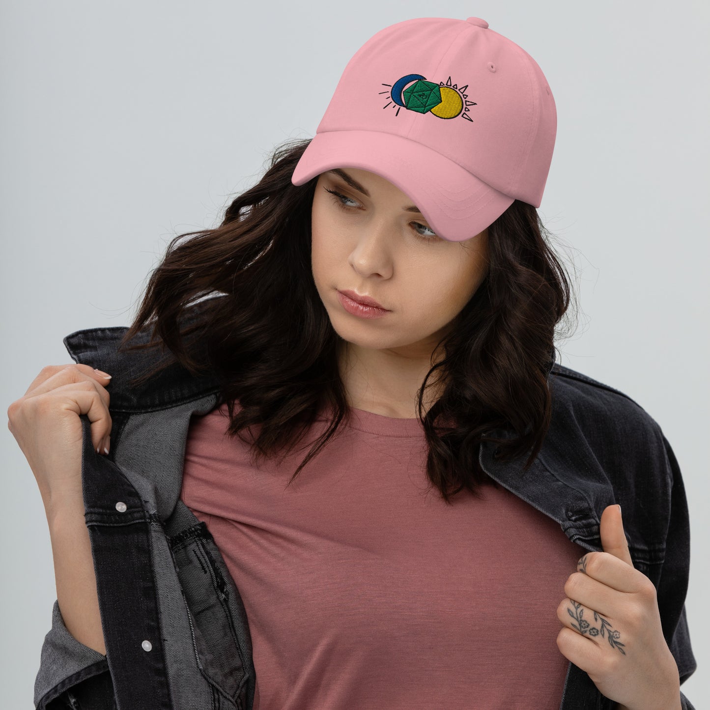 A model wears the Celestial D20 hat in pink, featuring a green D20 flanked by a blue moon and a yellow sun.