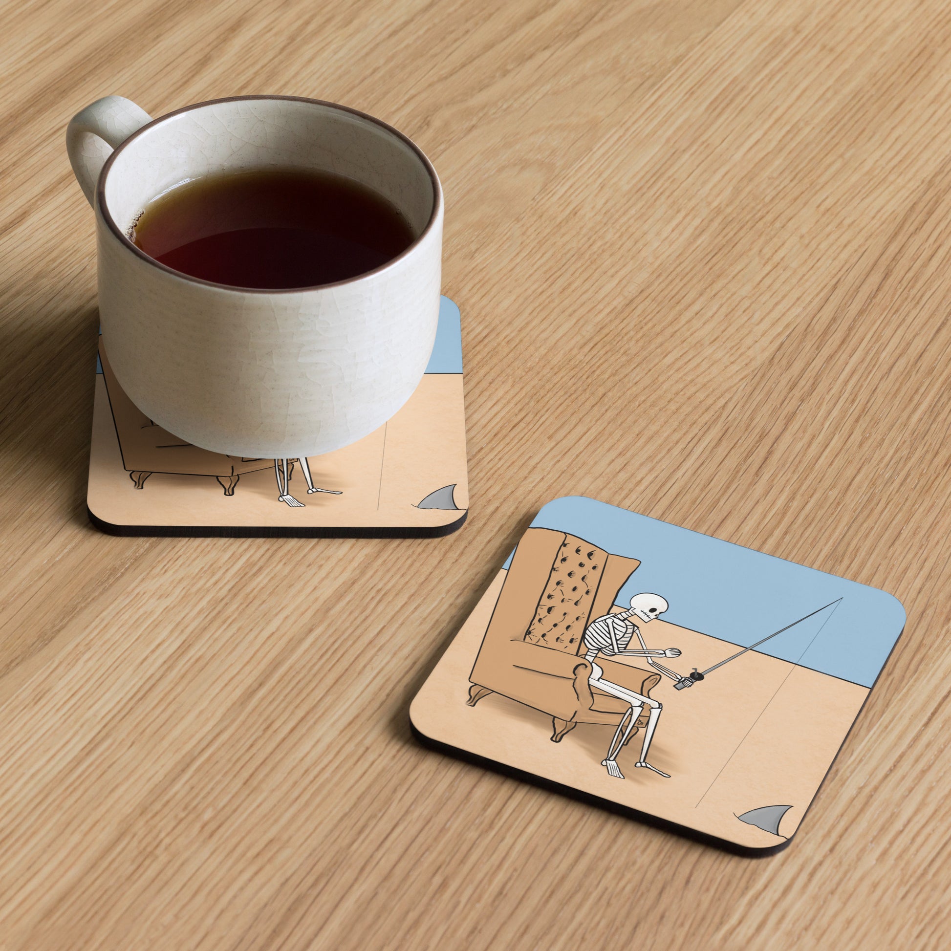 A pair of Dead Fishing coasters sit on a table, one holding a mug of liquid and the other uncovered to show the illustration of a skeleton sitting in a chair fishing in a sea of sand and a shark fin approaching.