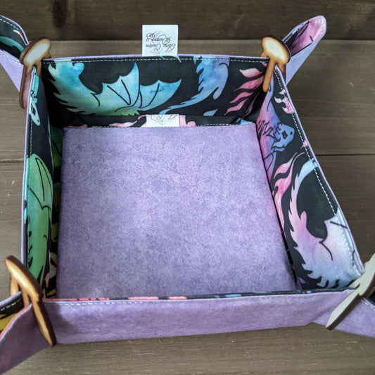 A dice tray with rainbow dragon fabric inside, purple fabric outside, a matching insert in the bottom, and wooden clips in the corners sits on a wood surface.