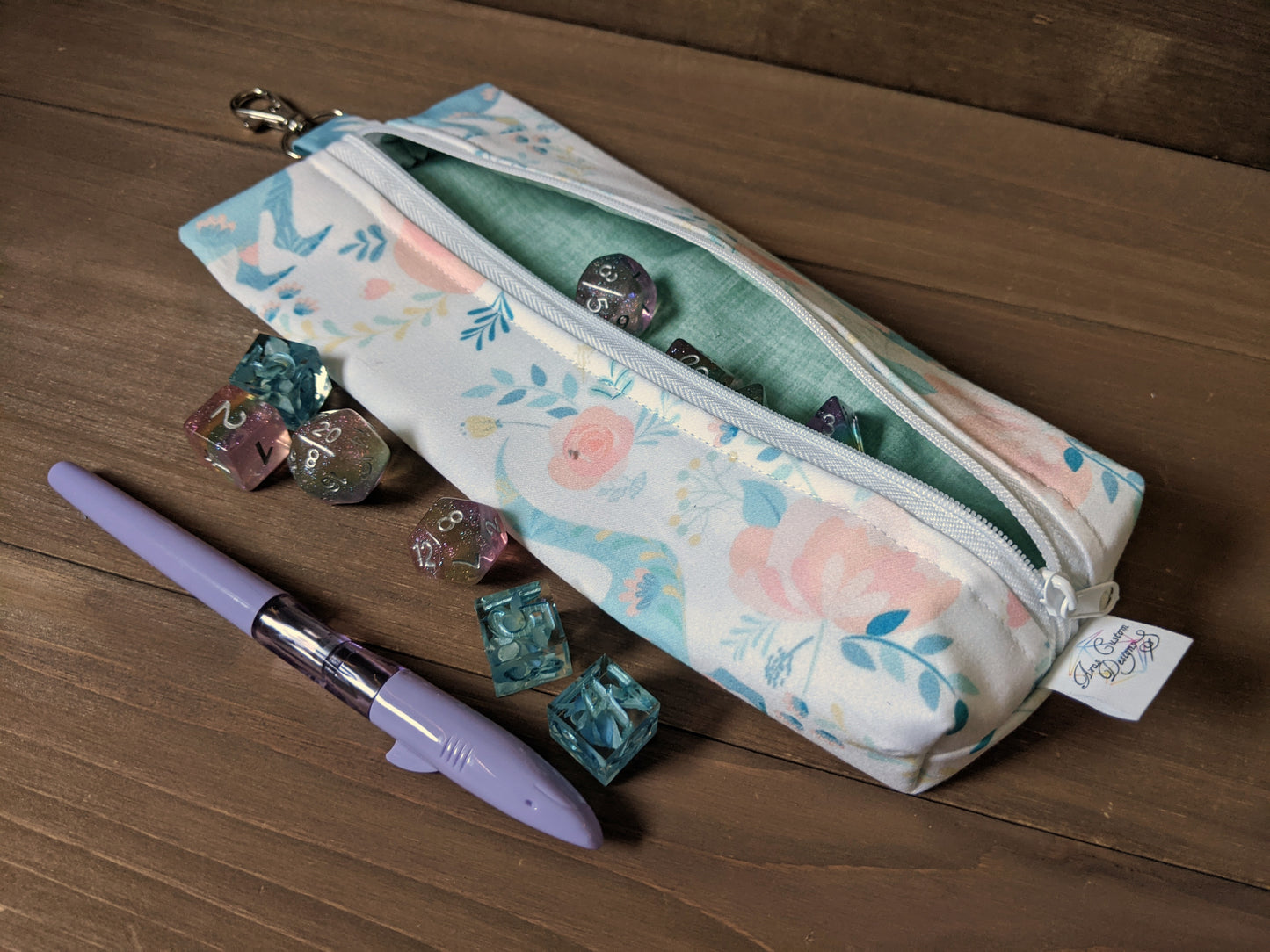 A zippered bag with pastel floral fabric featuring T-Rexes sits on a wood surface with dice and a shark pen.