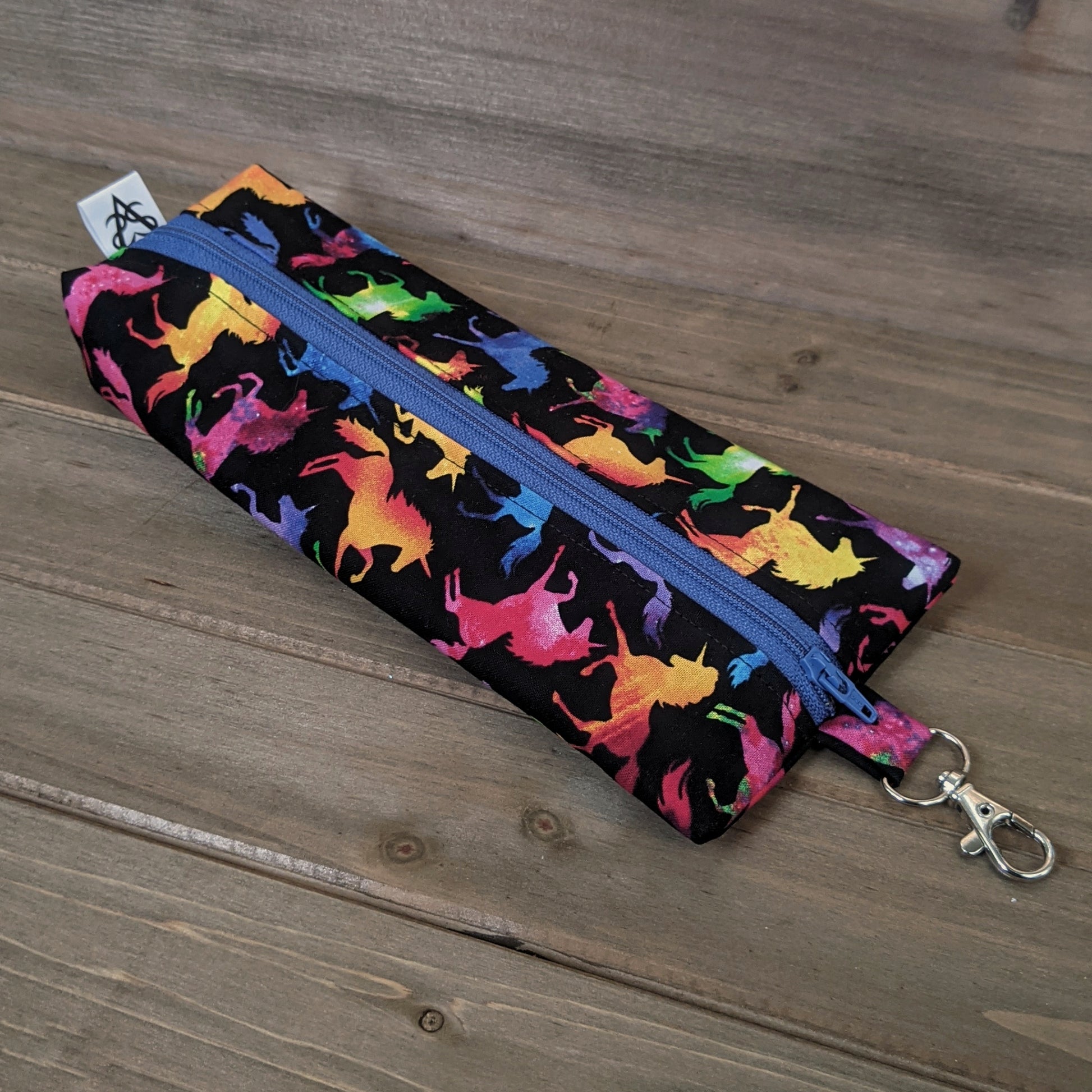 A zippered bag in the shape of a wedge with a tag at the bottom and a lobster clasp at the narrow end, made with rainbow galaxy unicorn fabric.