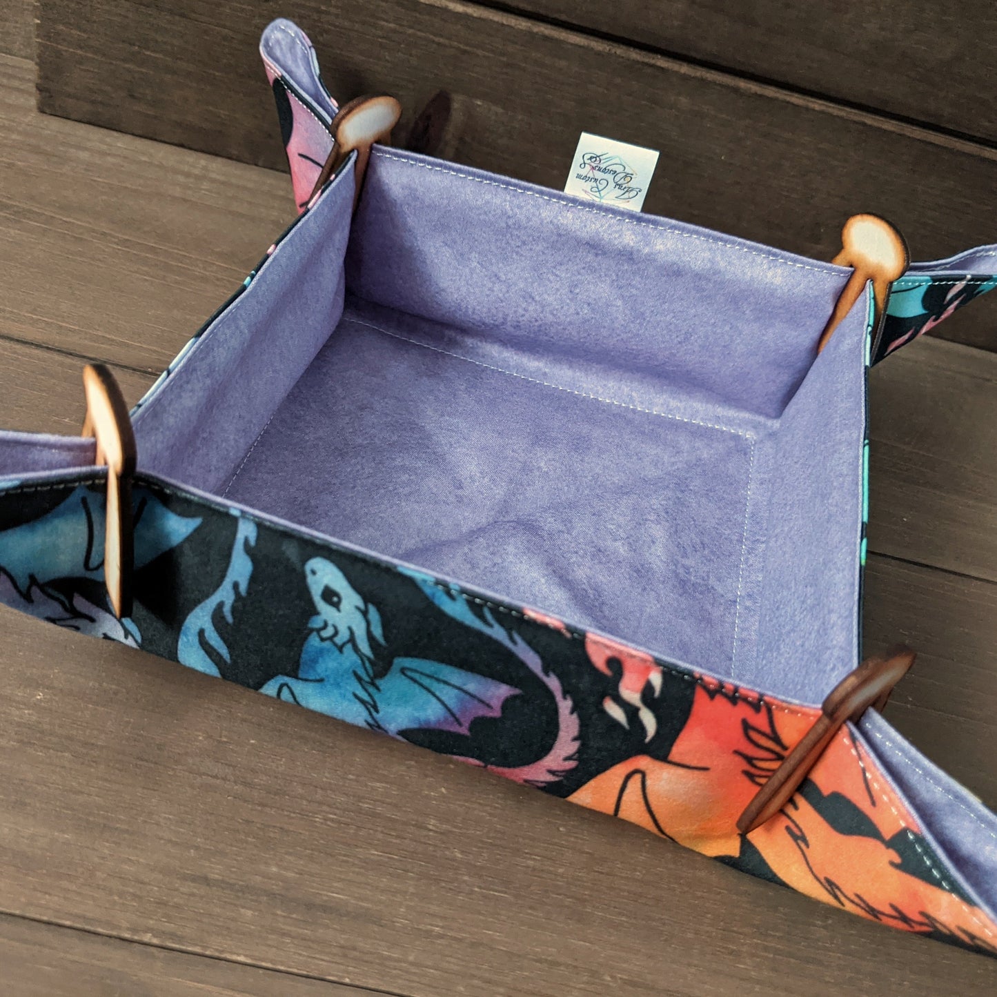 A dice tray with rainbow dragon fabric outside, purple fabric inside, and wooden clips in the corners sits on a wood surface.