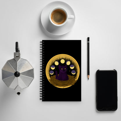 A black notebook with a painting of a dark purple cat with golden eyes sitting in a nest, ringed by the phases of the moon and golden stars behind it.